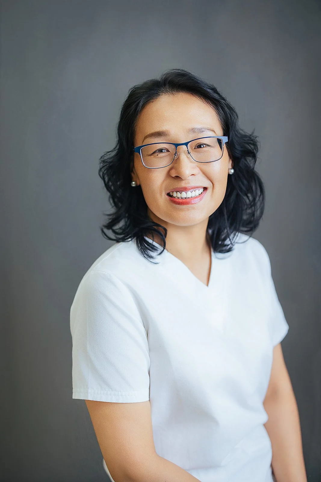 Durham Tech nursing student with short dark wavy hair and blue glasses, joyful smile in white scrubs, professional headshot in Durham NC