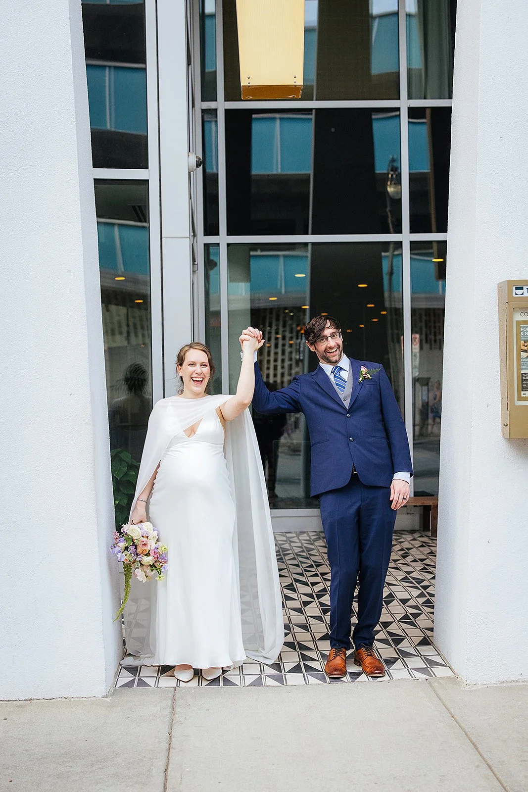 Newlywed couple raises their joined hands in celebration while standing in a modern building doorway with a graphic black and white tile floor, downtown Durham NC