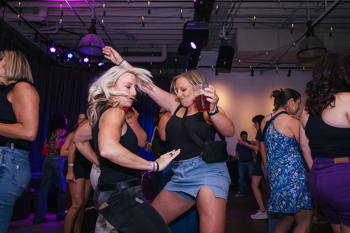 Two women dancing energetically at the Dance Her event in Raleigh — one with hair flying, one raising a beer — captured by a Raleigh event photographer
