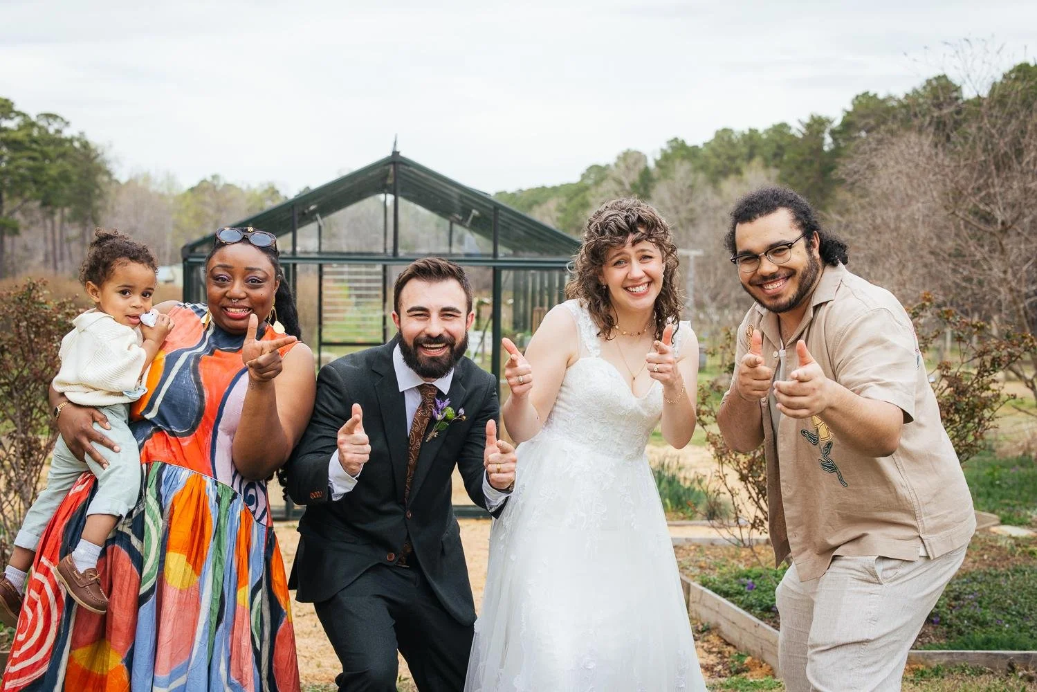 Bride, groom, and friends including an infant in a colorful carrier giving thumbs up outside the greenhouse at Honeysuckle Tea House Chapel Hill NC