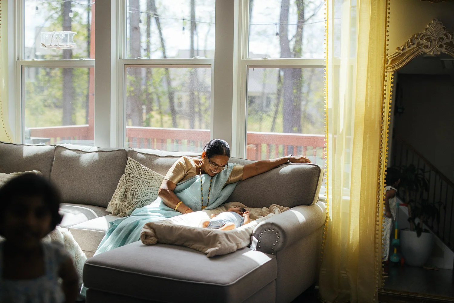Grandmother in a light blue saree sits on a large sofa with the newborn across her lap — golden yellow curtains and trees visible through large windows behind her — in-home newborn session in Apex, NC.