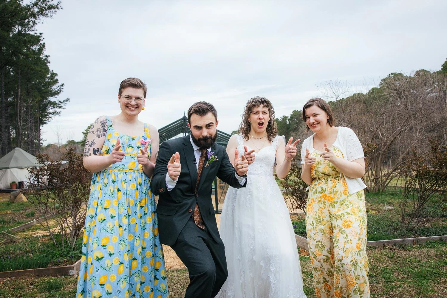 Bride Emily and groom Connor with two friends in floral and yellow floral jumpsuits laughing and pointing with playful energy outside the greenhouse at Honeysuckle Tea House Chapel Hill NC