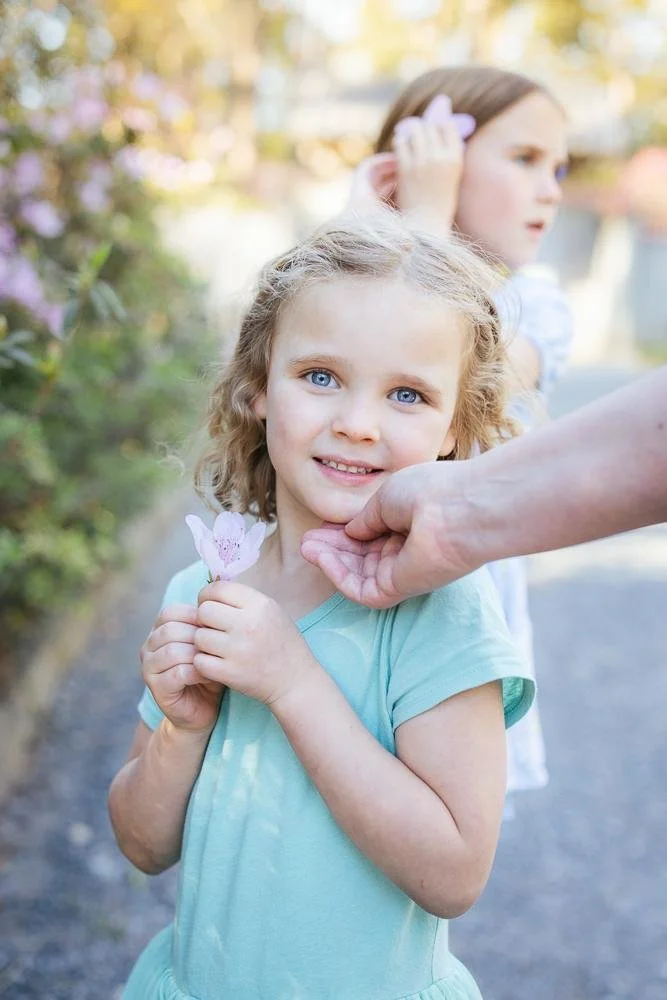 Young girl with bright blue eyes holding a pink azalea flower and smiling at the camera at Duke Gardens Durham NC — family photographer