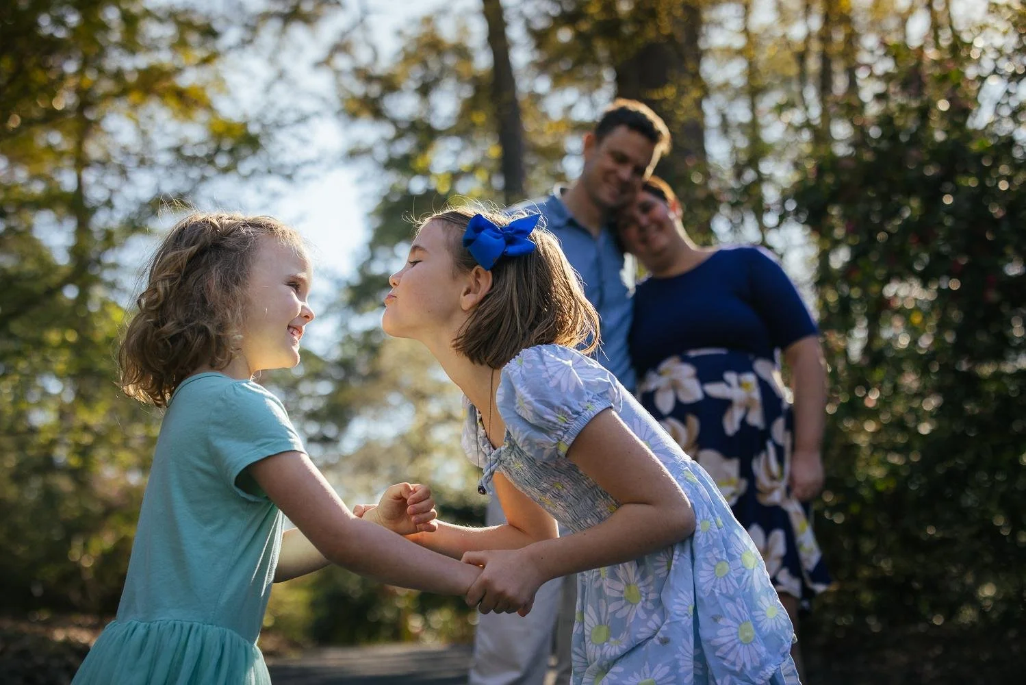 Two sisters holding hands and dancing together on a wooded path at Duke Gardens in Durham NC while parents watch in background