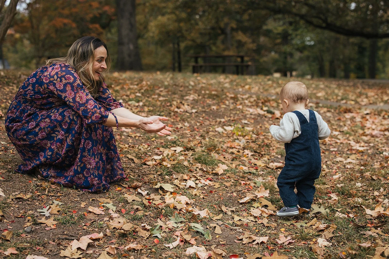 Mom crouches with arms wide open as baby boy walks toward her in fall leaves at Duke Park during Durham NC first birthday photography session