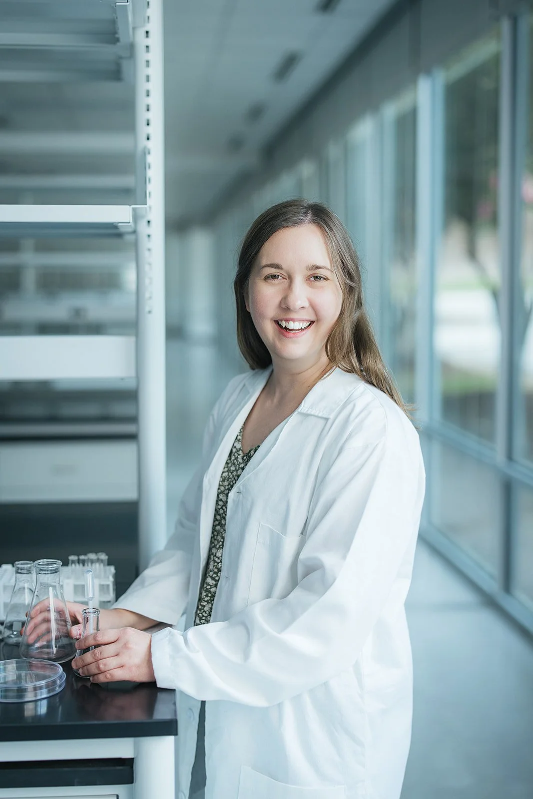 WomanLongBrownHairWhiteLabCoatSmilingHoldingBeaker