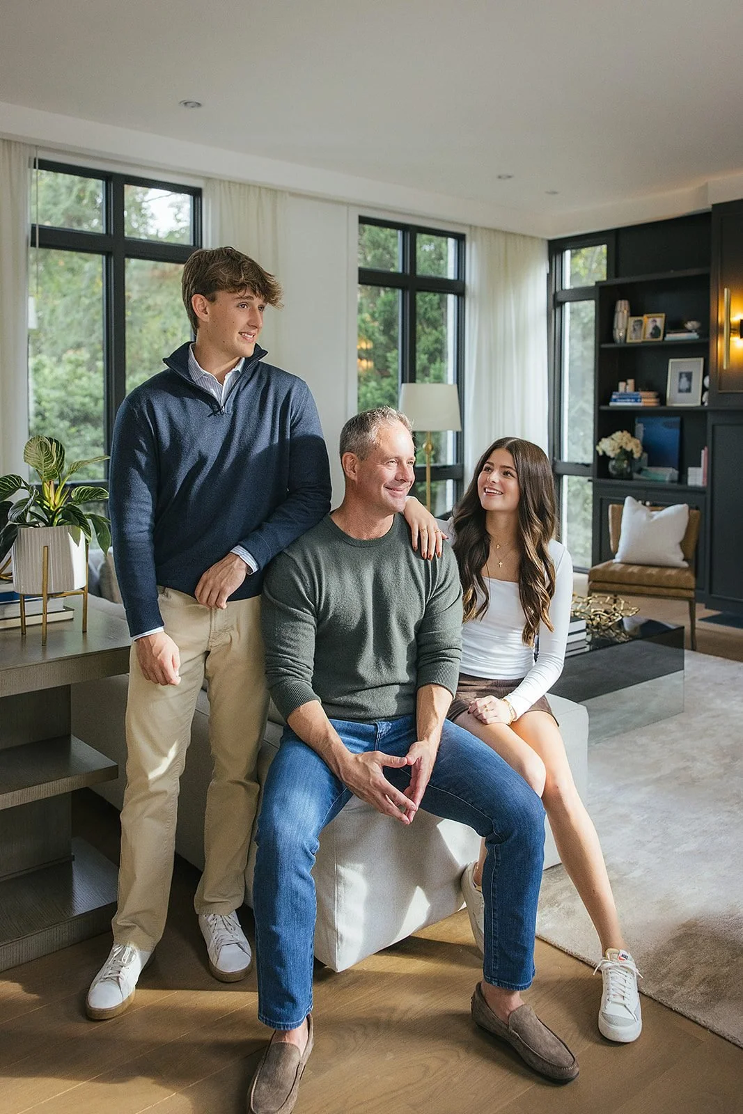 A father and two teenagers laugh together in a bright, modern living room during their in-home family session in downtown Raleigh, NC — son standing on the left, dad in the center, daughter perched on an ottoman to the right.