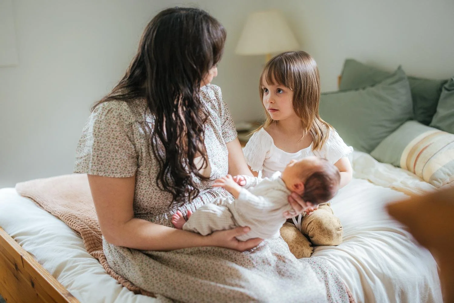 Mom holding newborn baby on bed while toddler girl sits beside her looking at her baby brother — Durham NC newborn photographer