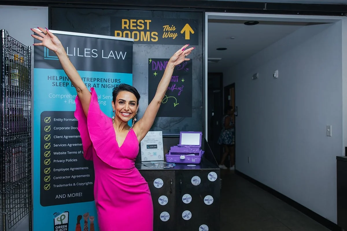 Dance Her organizer in a hot pink dress posing with arms raised in front of the Dance Her sign and sponsor table at Transfer Co Food Hall in Raleigh