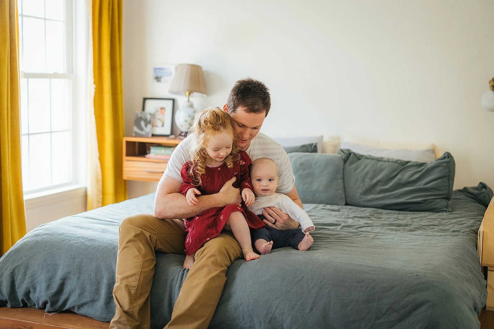 Dad holds both toddler and baby on bed in a tender moment during Durham lifestyle family photography session