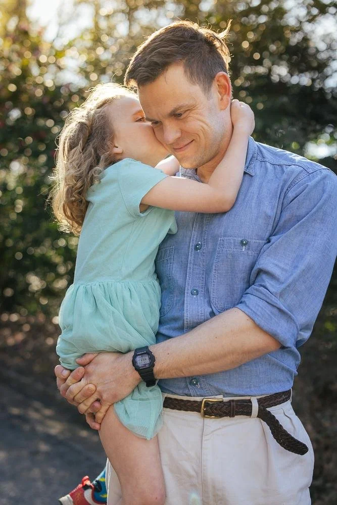 Young daughter giving her dad a sweet kiss on the cheek while he holds her during a Duke Gardens family session in Durham NC