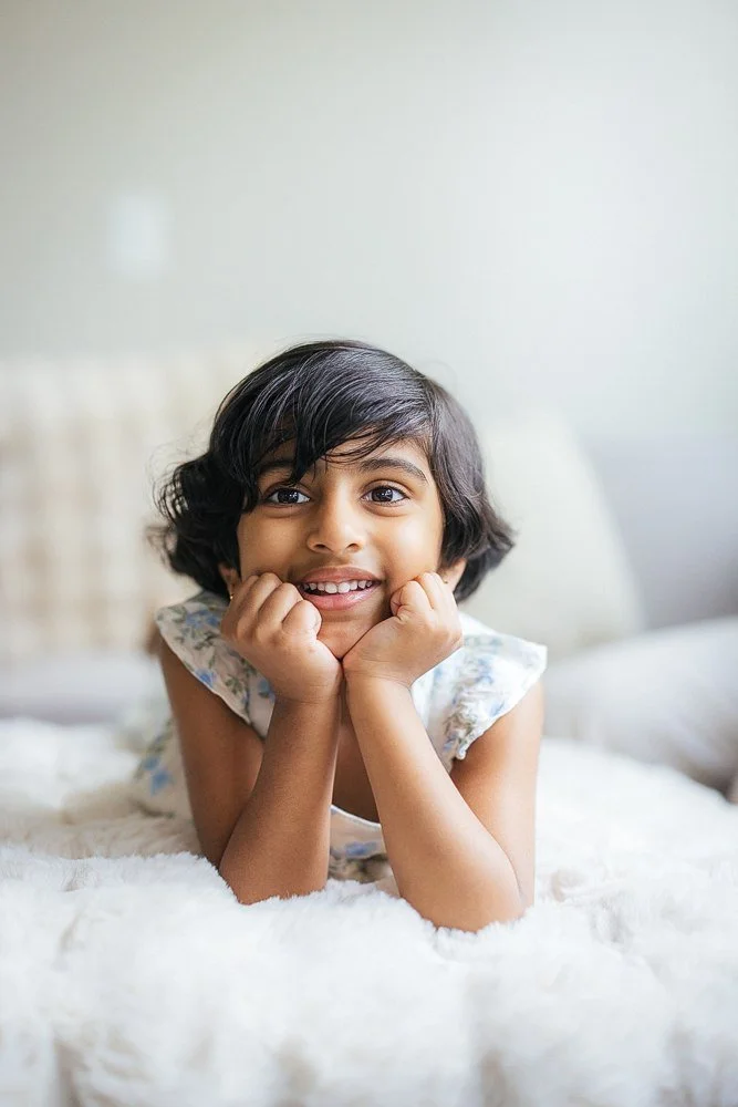 Young girl with dark wavy hair rests her chin in both hands on a fluffy white bed, smiling directly at the camera — big sister portrait from an in-home newborn family session in Apex, NC.
