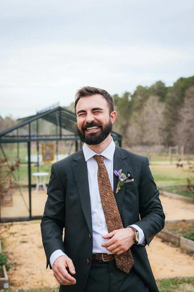 Groom Connor laughing broadly in his dark green suit and brown paisley tie — boutonniere with purple flowers — portrait at Honeysuckle Tea House Chapel Hill NC wedding