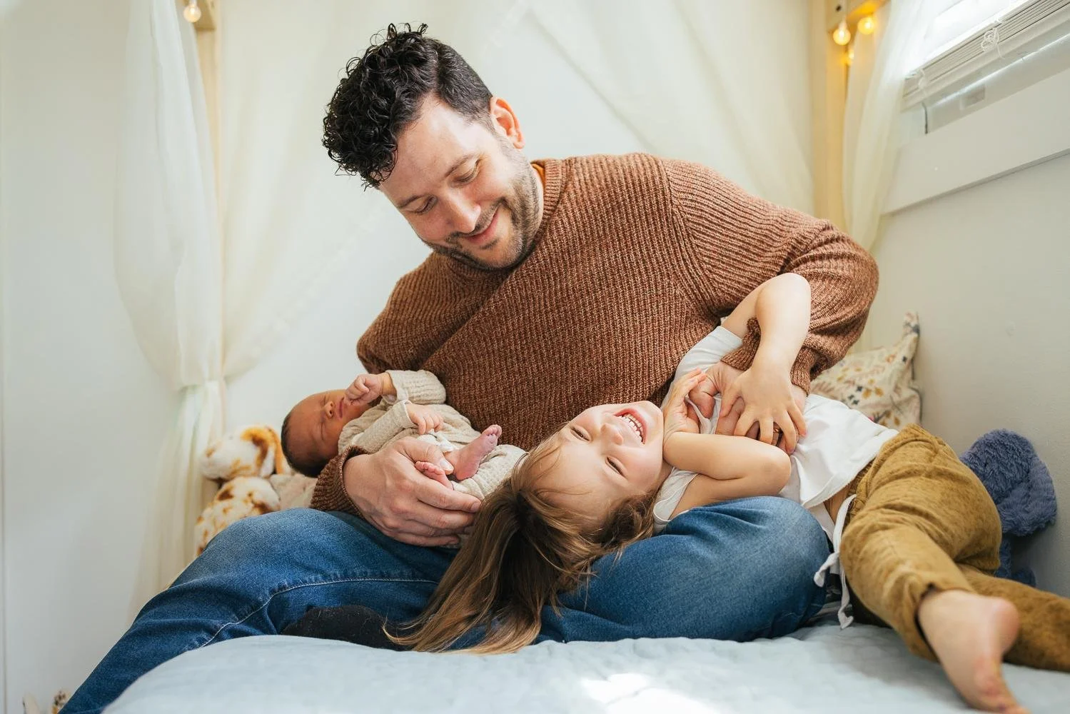 Dad in a brown sweater holding his newborn baby while his toddler daughter laughs beside him — Durham NC newborn photography