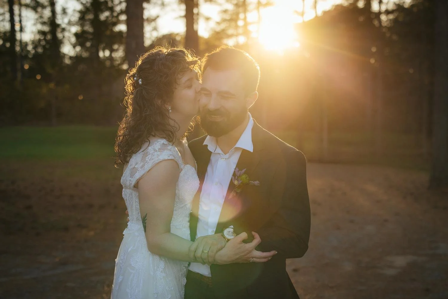 Bride kissing groom on the cheek at golden hour sunset — warm glowing light through pine trees at Honeysuckle Tea House Chapel Hill NC wedding portraits