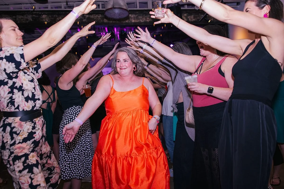 Woman in a bright orange gown walking through a tunnel of raised arms from cheering women at the Dance Her popup, Transfer Co, Raleigh NC