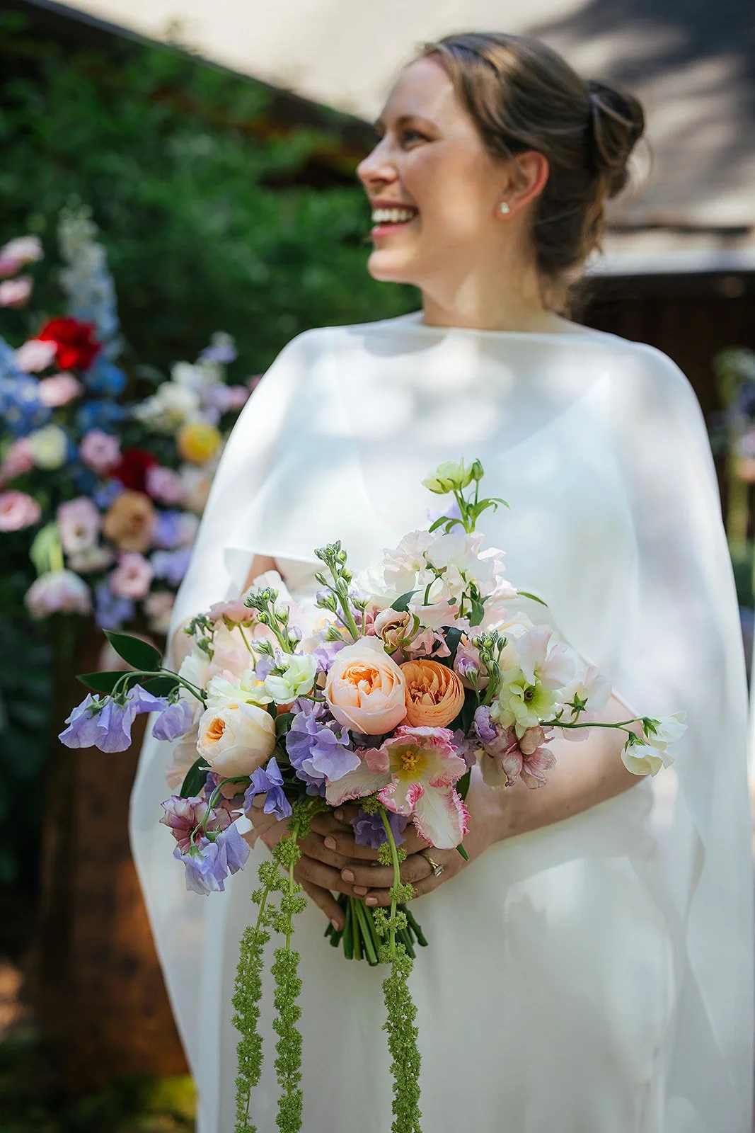 Pregnant bride in a white cape dress holds a lush pastel bouquet of garden roses, sweet peas, and lavender while laughing outdoors, Durham NC wedding