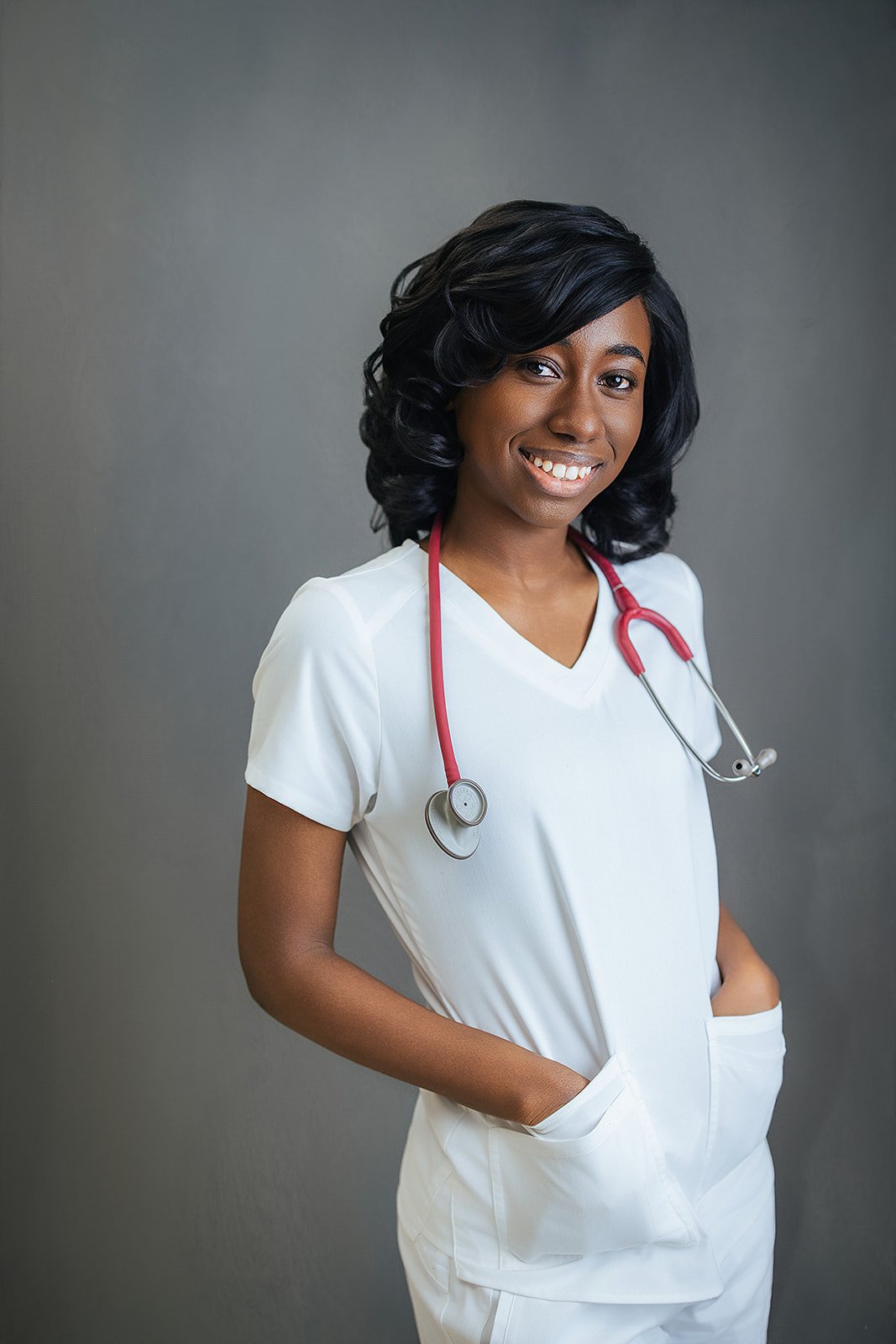 Durham Tech nursing student with curly black hair, hands in pockets, beaming smile in white scrubs and red stethoscope, professional headshot Durham NC