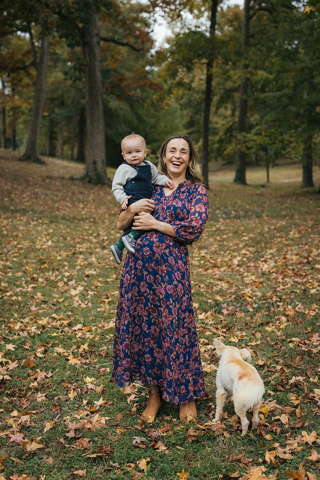 Mom stands laughing with her one - year - old baby boy and white fluffy dog among fall leaves at Duke Park during Durham NC motherhood session