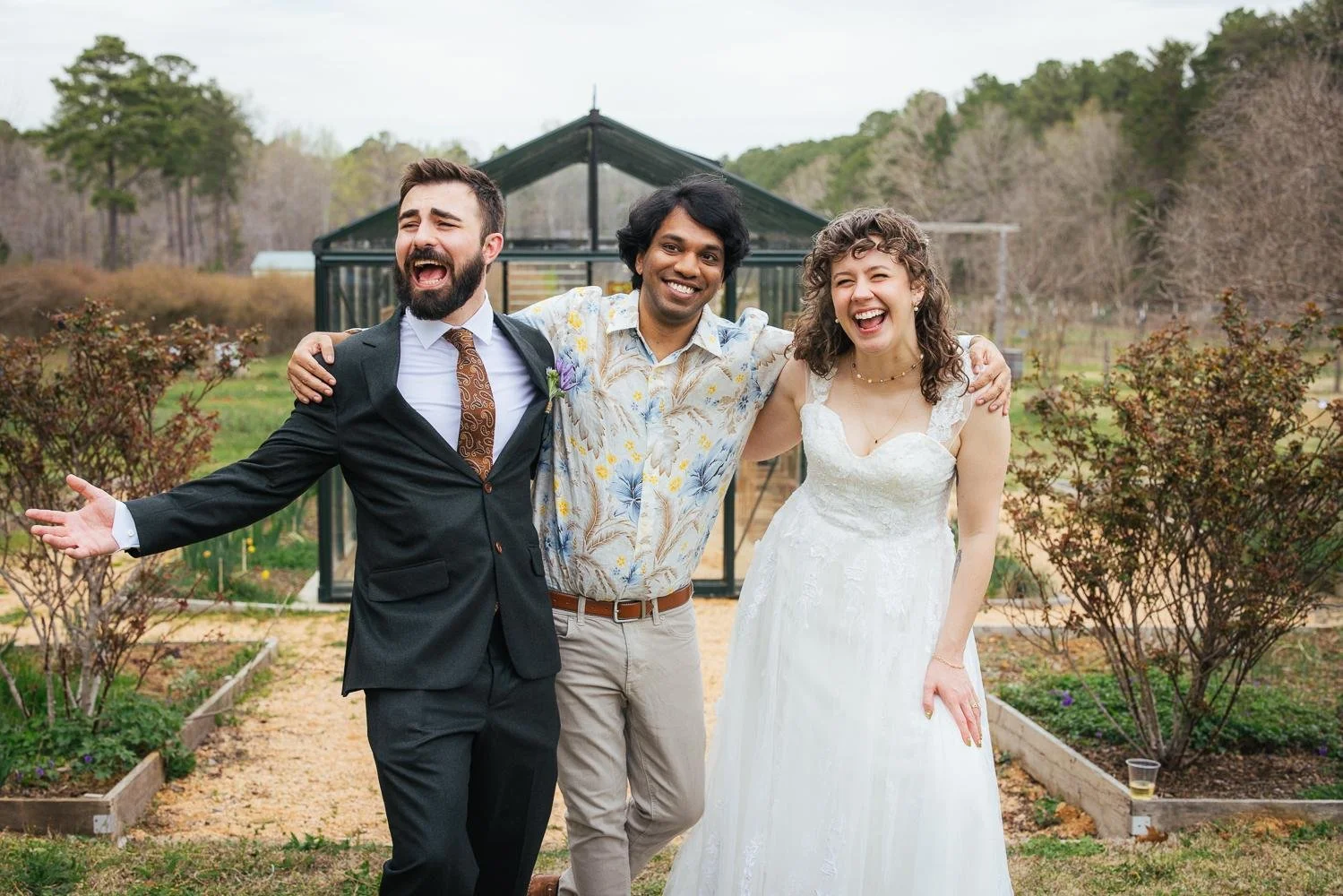 Groom with arms wide open laughing alongside a friend in a patterned shirt in the garden at Honeysuckle Tea House Chapel Hill NC wedding