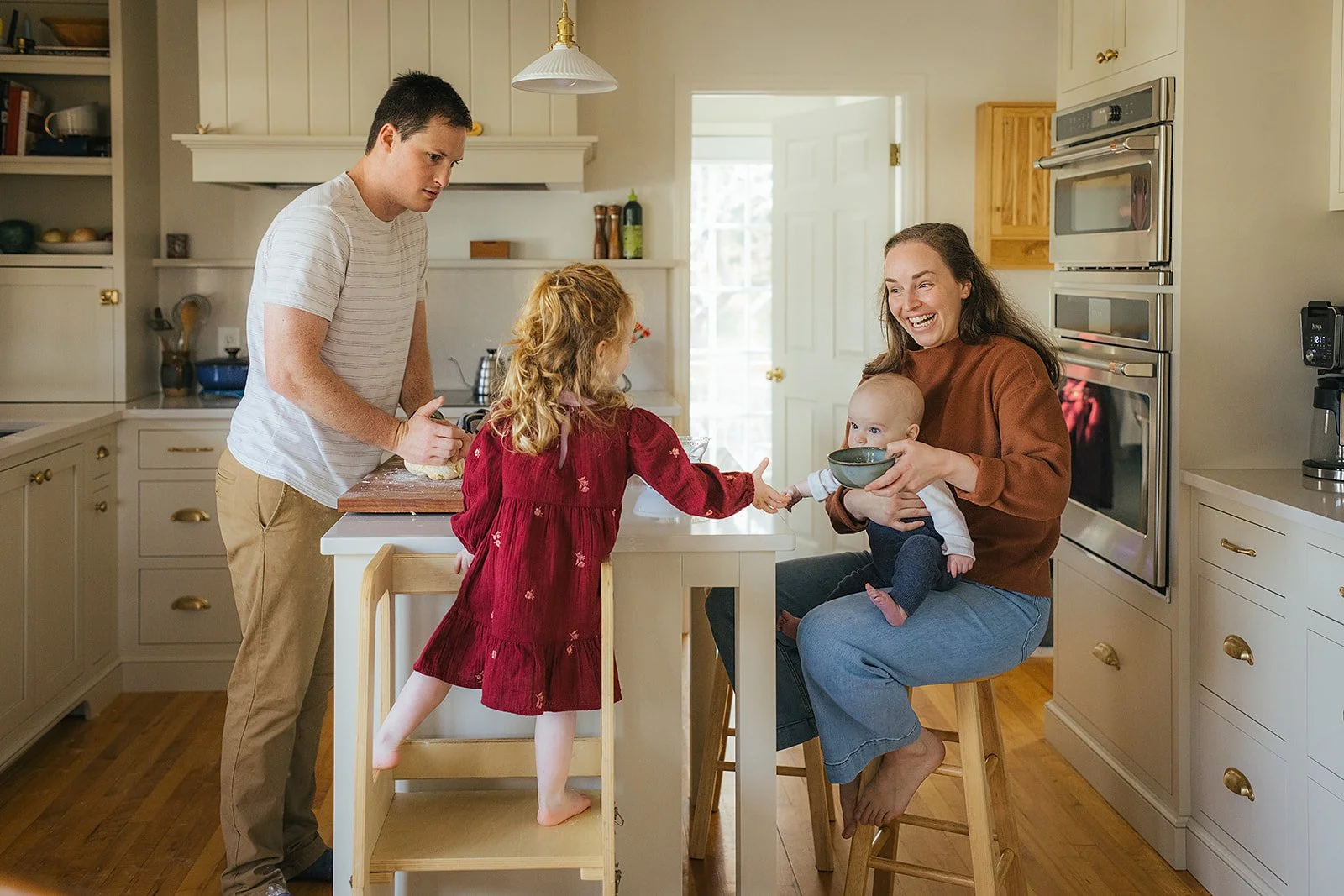 Family of four cooking together in kitchen — toddler on learning tower helping dad while mom holds baby during Durham lifestyle session