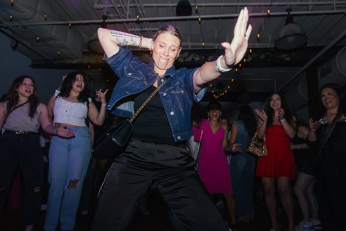 Woman in a denim jacket throwing a big dance move on the floor — one hand behind her head, one arm outstretched — as the crowd cheers at the Raleigh Dance Her event