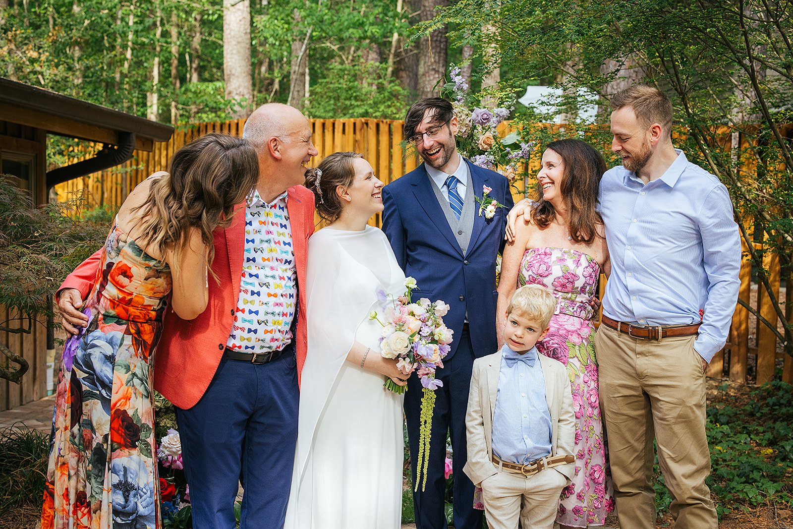Newlywed couple surrounded by joyful, colorfully dressed family members of all ages laughing together in a wooded backyard, Durham NC wedding