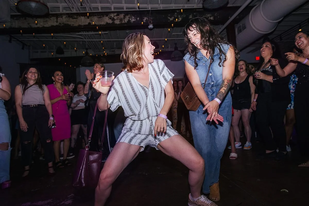 Two women cracking up while dancing together at the Dance Her popup in Raleigh, one in a striped romper holding a pink drink