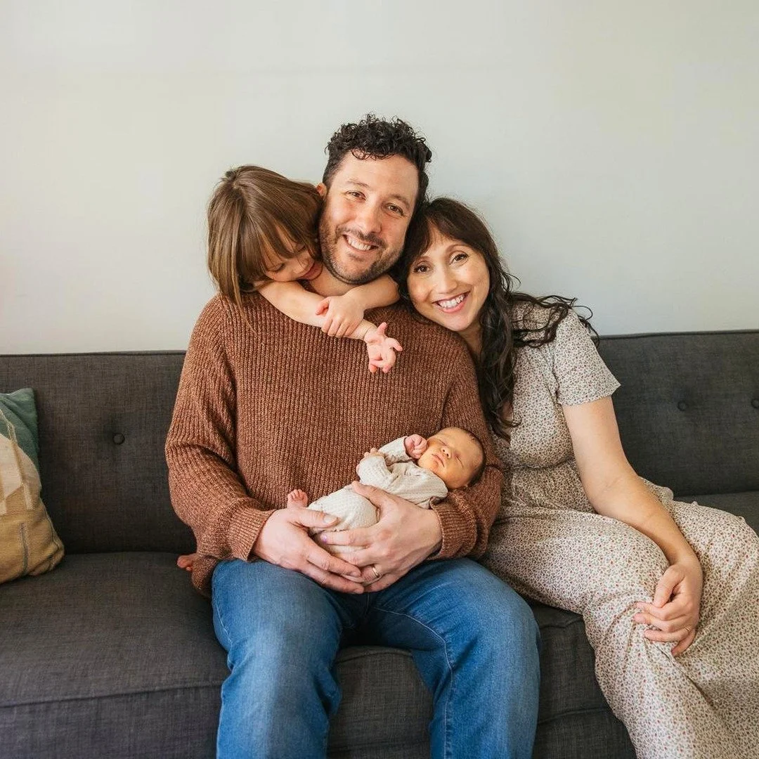 Family of four smiles together on a couch during an in home newborn session in Durham NC