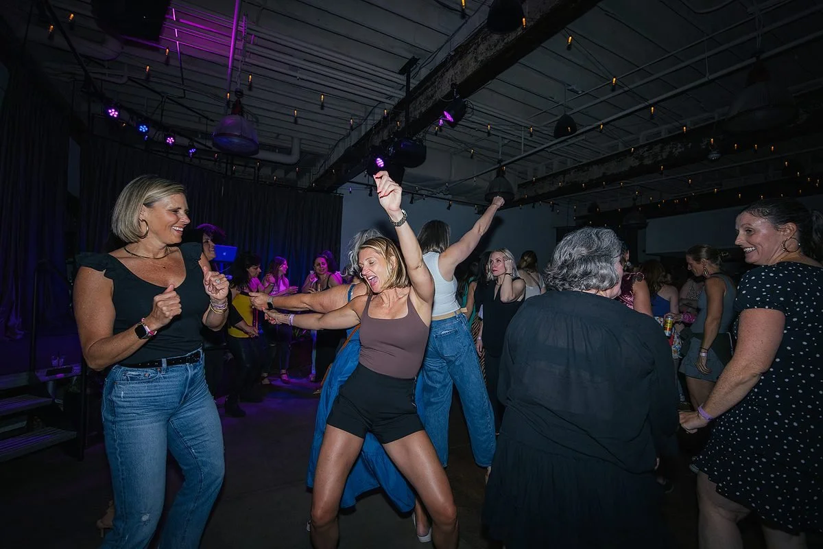 Wide dance floor shot at Transfer Co Raleigh — woman in the center with arms raised, laughing, surrounded by women of all ages dancing freely