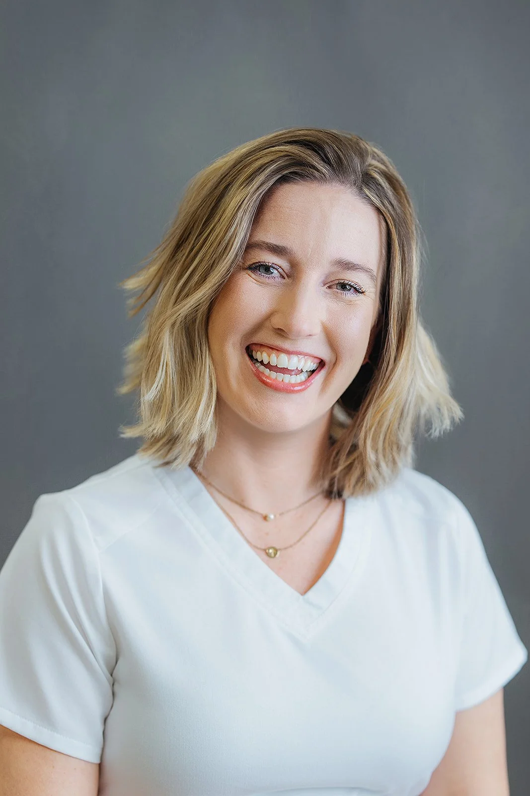 Durham Tech nursing student with short blonde hair laughing joyfully in white scrubs with layered necklaces, professional headshot in Durham NC