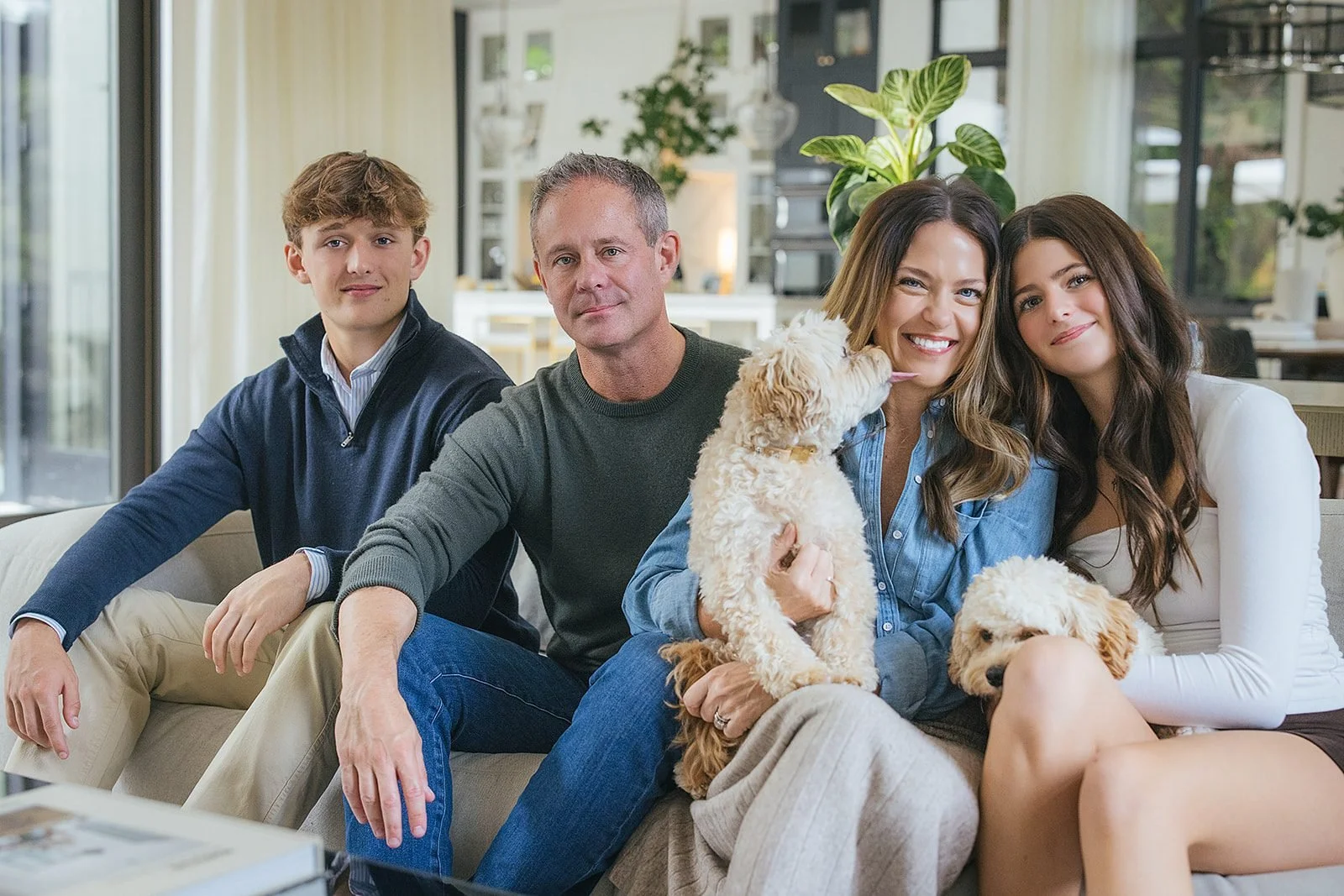 A family of four sits together on a cream sofa smiling at the camera, their fluffy goldendoodle settled happily in mom's lap, captured during their in-home family session in downtown Raleigh, NC.