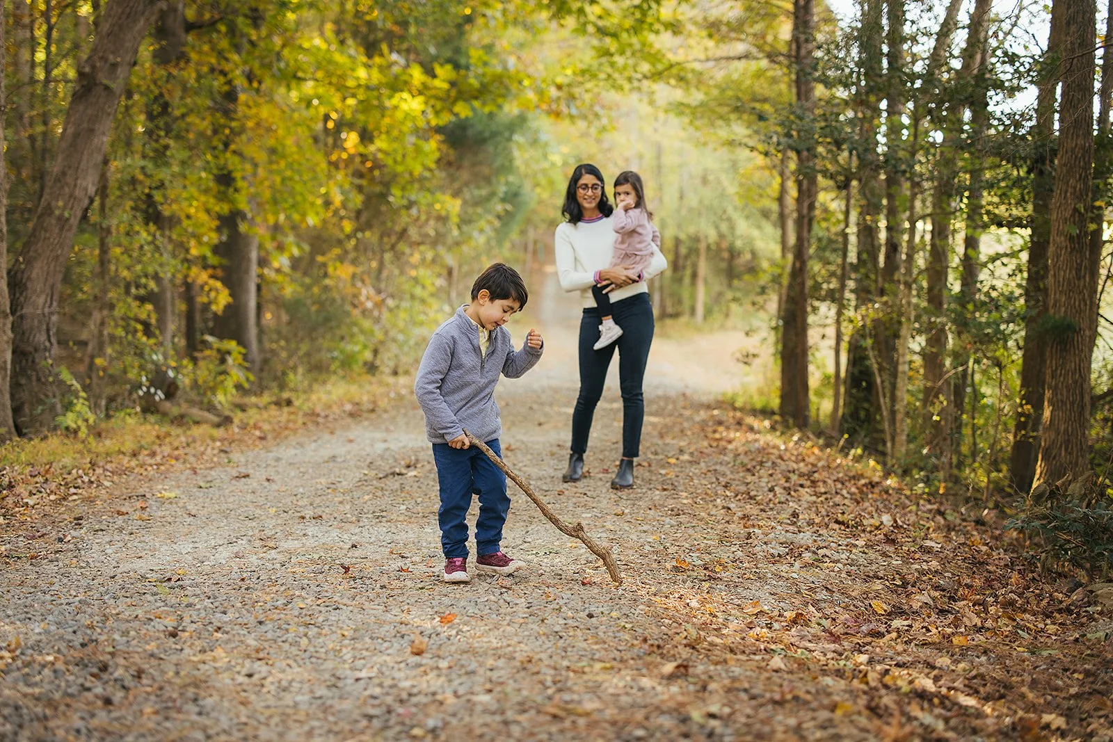Mom holding her daughter while her son plays with a stick on a wooded path at Blackwood Farm Park, natural light filtering through the trees