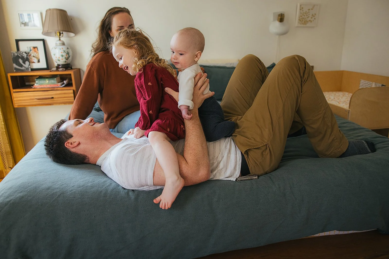 Dad plays with toddler and baby on bed while mom watches during Durham in-home family photography session