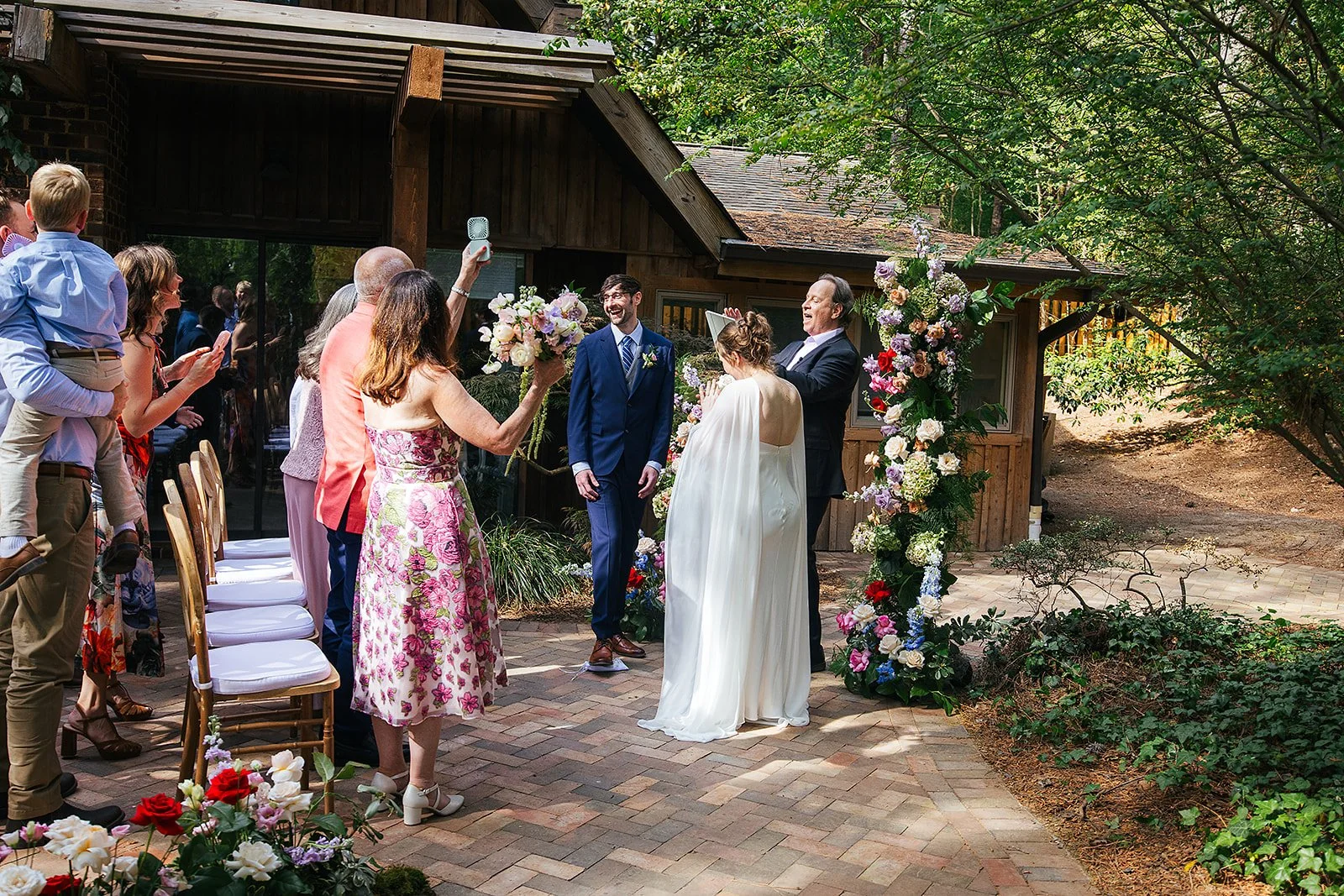 Guests cheer and photograph as the newlywed couple walks joyfully back through their intimate outdoor ceremony space, Durham NC wedding recessional