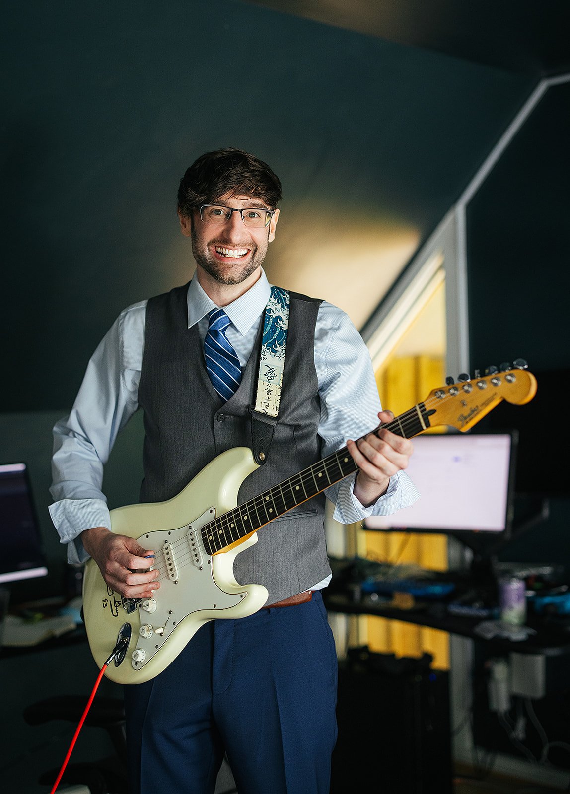 Groom in a gray vest and blue pants holds a cream Fender Stratocaster electric guitar and smiles broadly while getting ready in a music studio, Durham NC wedding