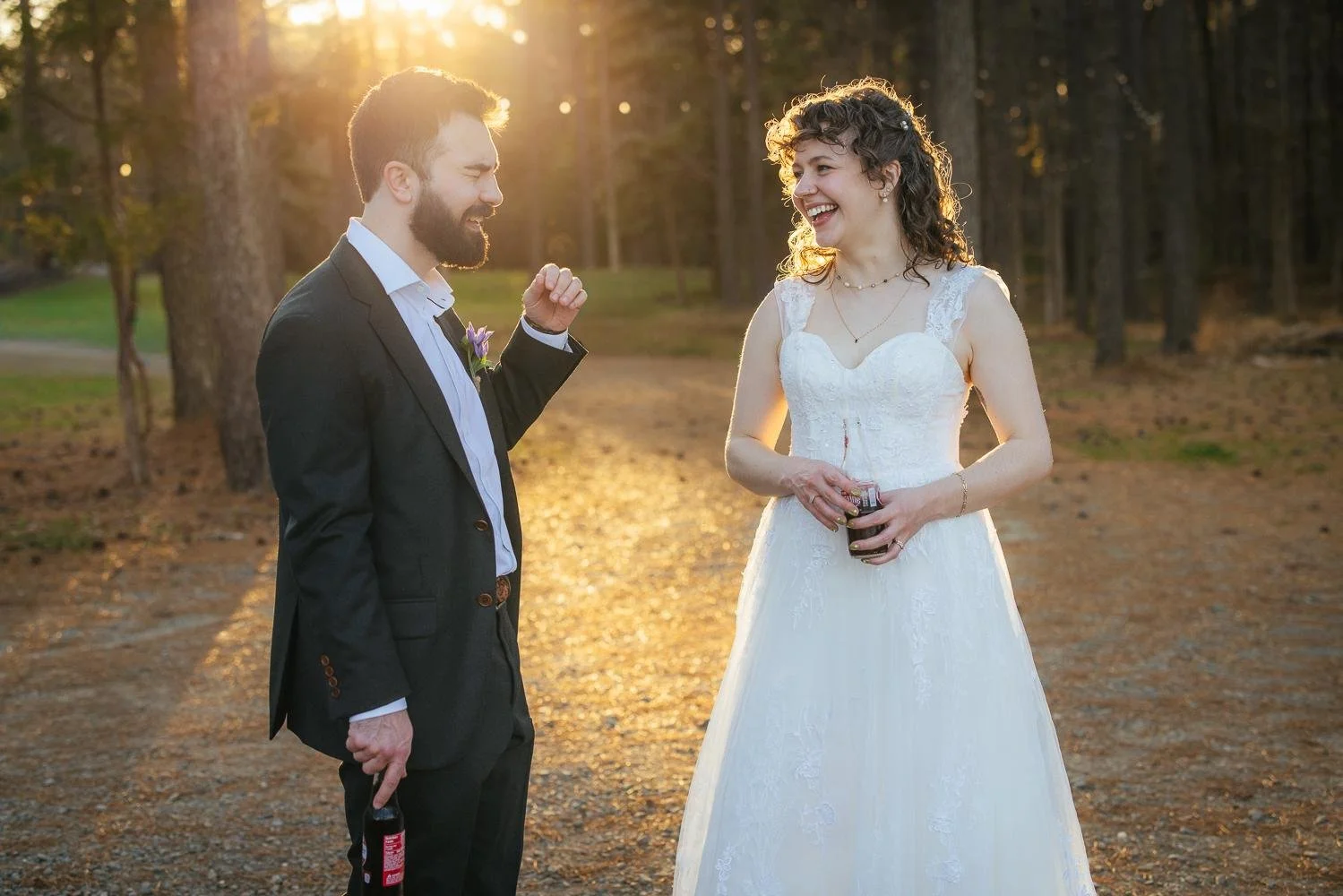 Bride and groom walking and laughing together on the pine path at sunset — each holding a drink — golden hour portraits at Honeysuckle Tea House Chapel Hill NC wedding