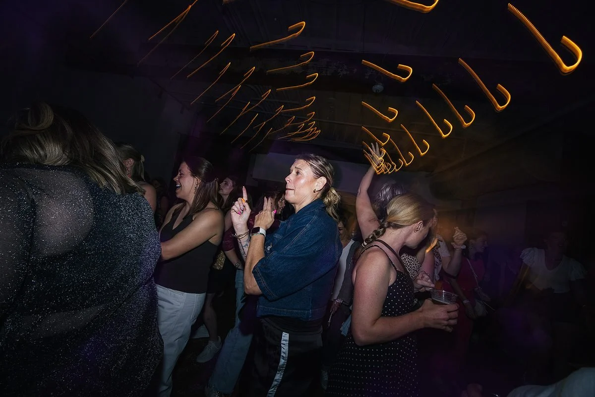 Crowd of women dancing at Transfer Co with glowing orange light trails arching overhead, captured with intentional motion blur at the Dance Her Raleigh event