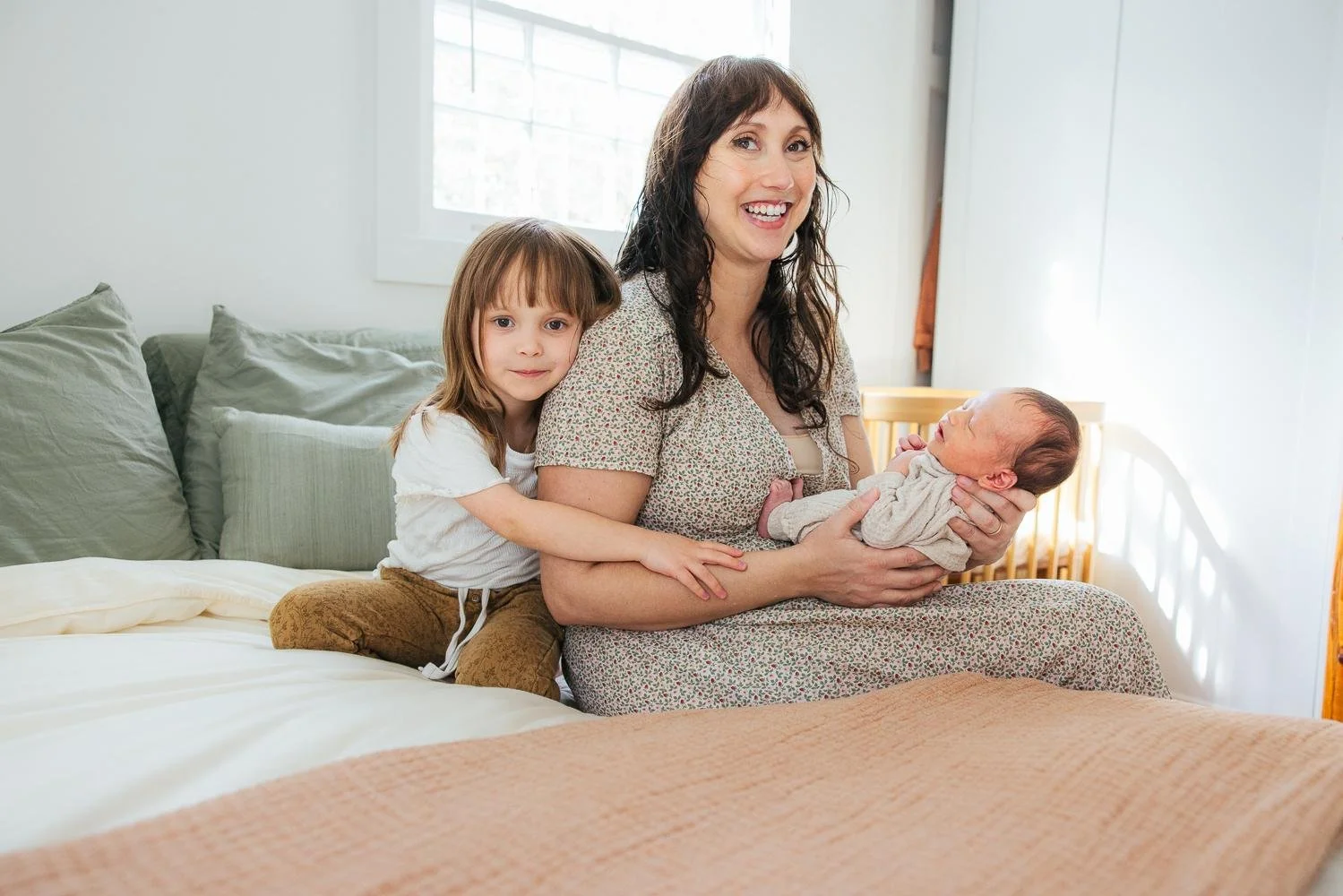 Mom sitting on bed holding newborn while toddler girl hugs her arm — all three looking at camera during Durham NC newborn session