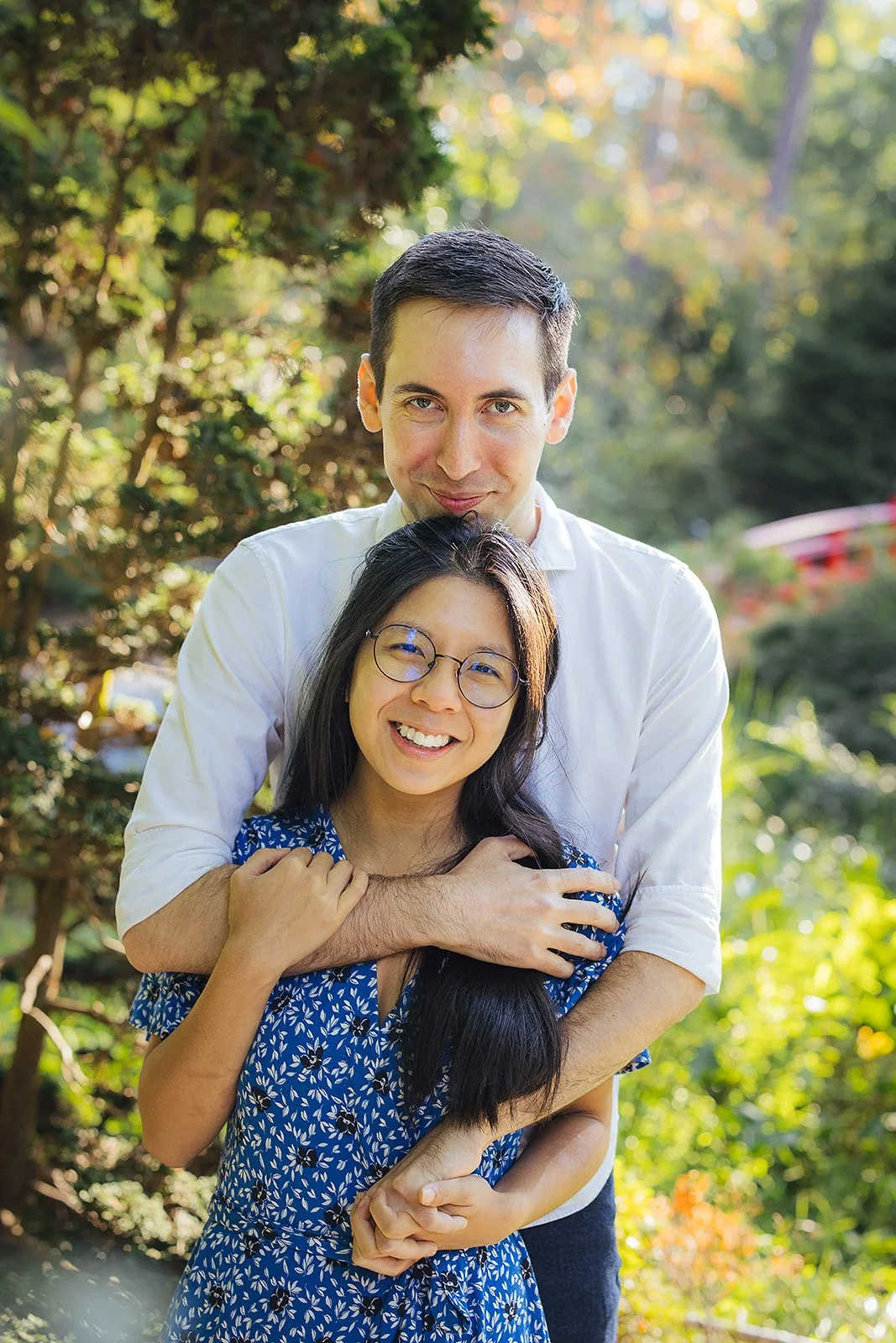 A couple hugs and hold hands surrounded by lush foliage after their surprise engagement during a photoshoot at Duke Gardens in Durham, NC