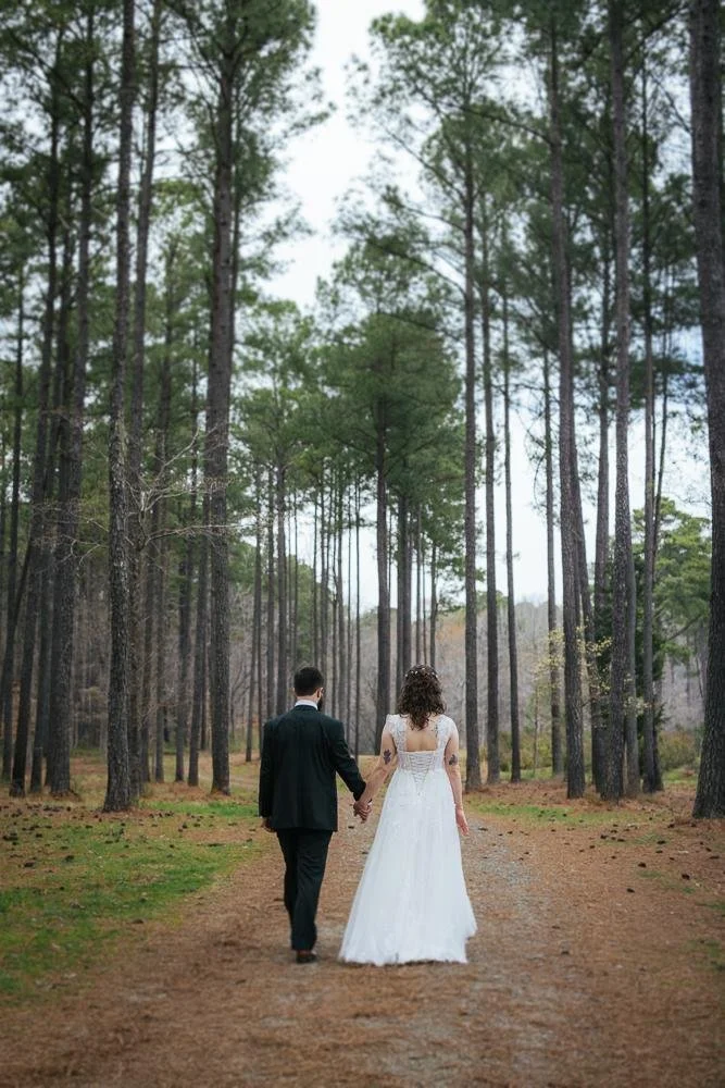 Bride and groom walking hand-in-hand away from camera down a pine tree-lined path at their Honeysuckle Tea House wedding in Chapel Hill NC