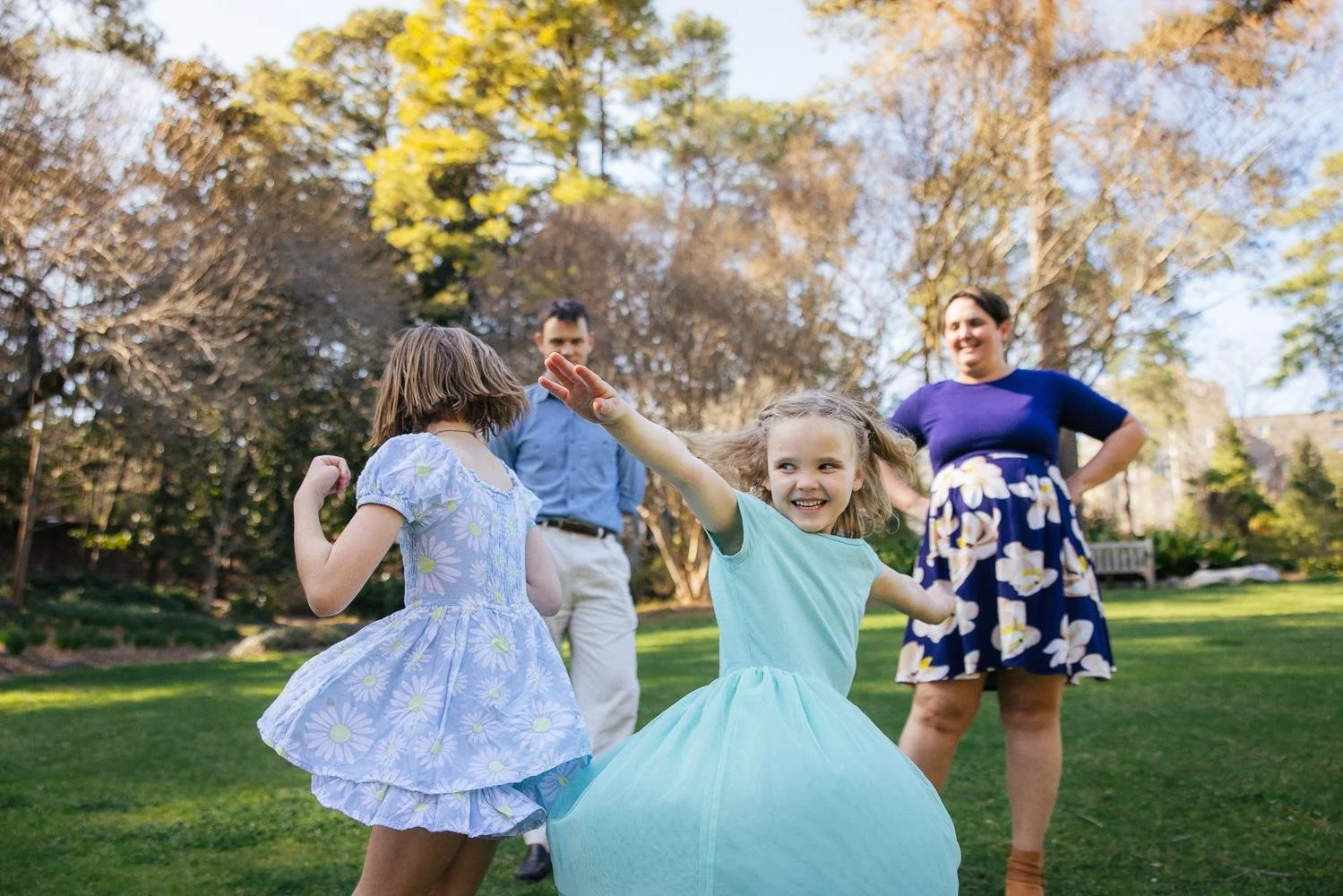 Family of four dancing and spinning on the green lawn at Duke Gardens during a Durham NC family photography session — mom, dad, and two daughters