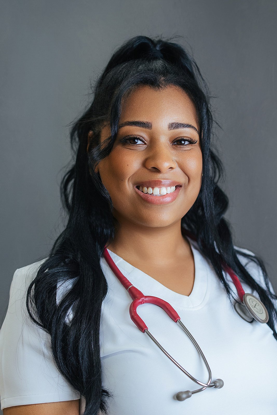 Durham Tech nursing student with long dark wavy hair, smiling warmly in white scrubs with red stethoscope, professional headshot in Durham NC