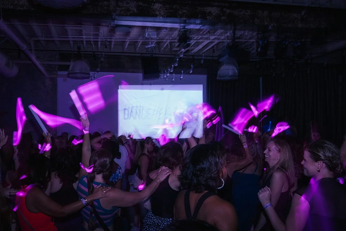 Full crowd waving pink glow sticks at the Dance Her popup in Raleigh, with the Dance Her logo projected on screen in the background at Transfer Co