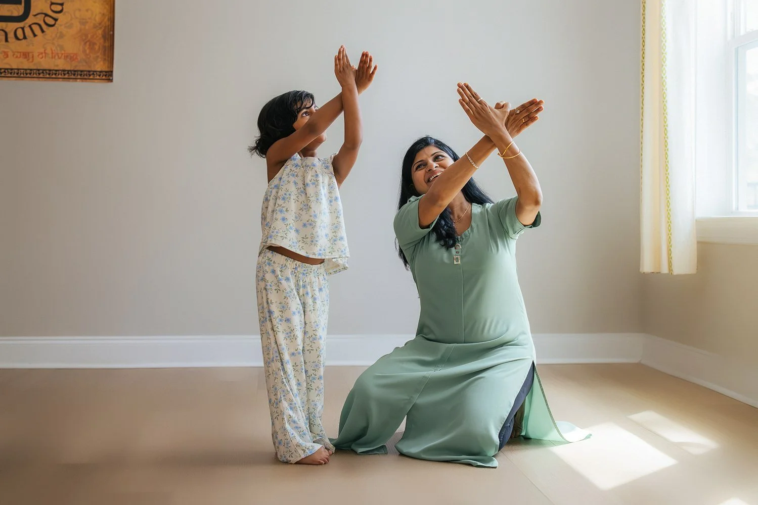 Mother in a sage green dress and her young daughter both raise their arms together in Indian classical dance gestures in a sunlit room — in-home family session in Apex, NC.