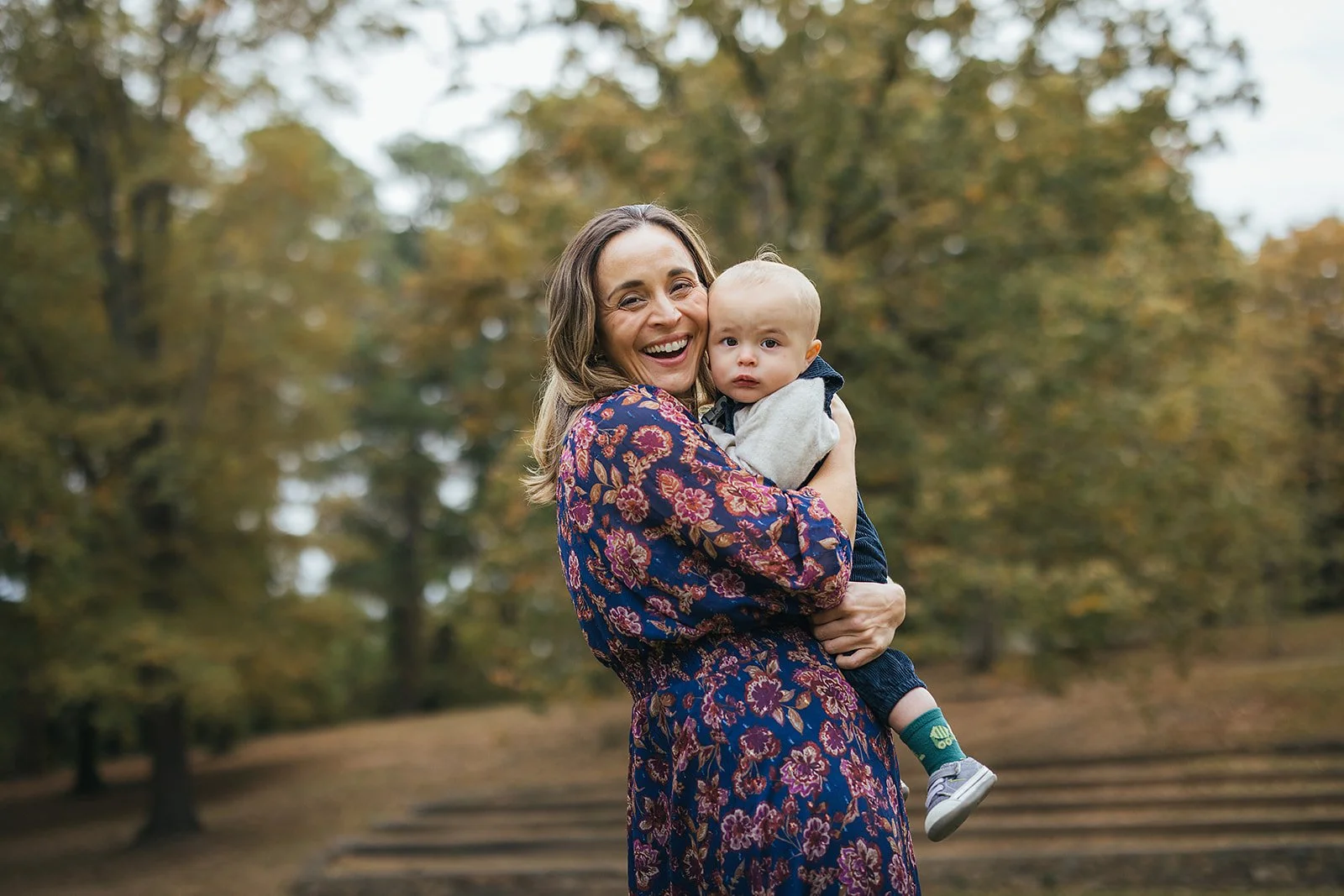 Mom laughs holding her one - year - old baby boy at Duke Park with fall foliage and the stone steps in the background in Durham NC