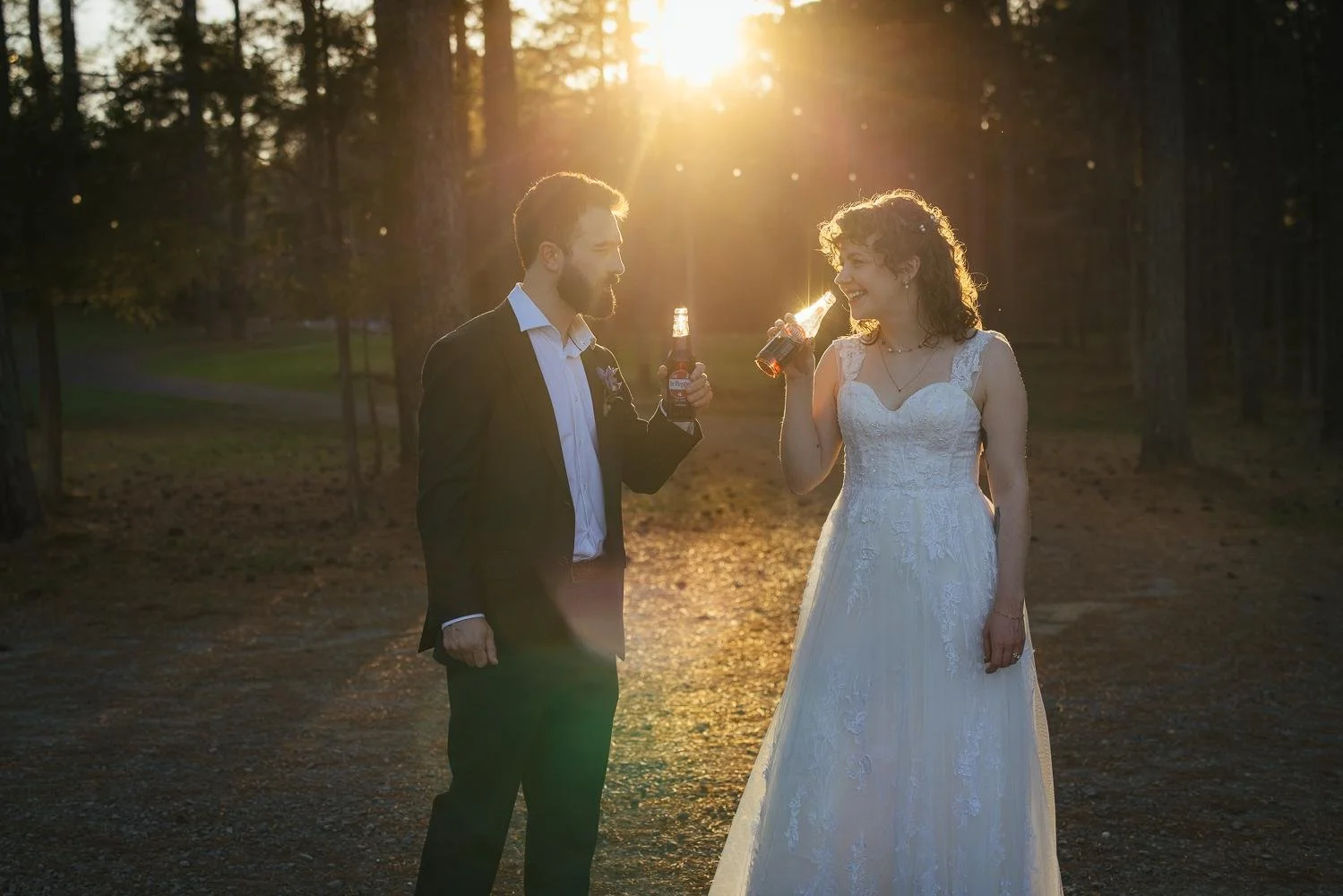 Bride and groom toasting with beer bottles at golden hour — gorgeous sun flare through the pine trees at Honeysuckle Tea House wedding Chapel Hill NC