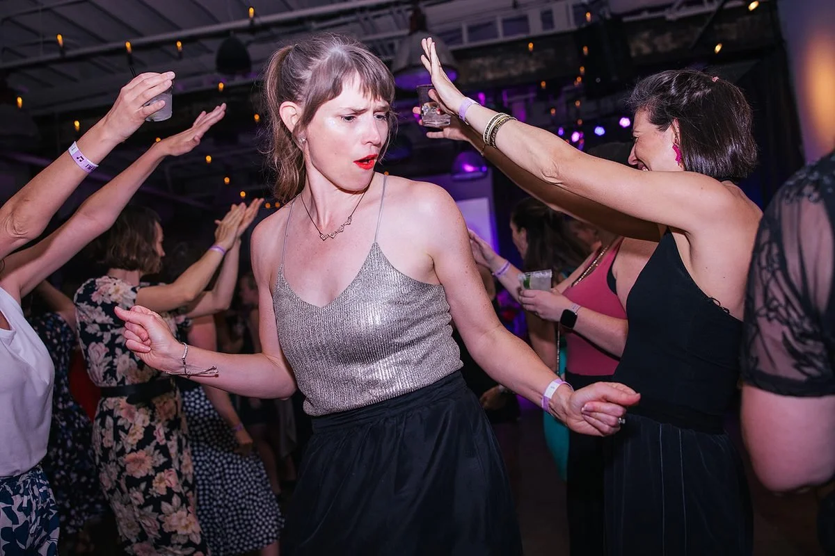 Woman in a silver top dancing in the middle of the floor at the Dance Her event in Raleigh, surrounded by other women with hands reaching in