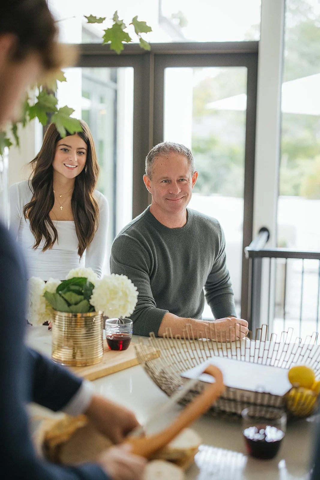 A father and daughter share a genuine laugh at the kitchen counter while someone prepares food in the foreground, a candid moment from their in-home family session in downtown Raleigh, NC.