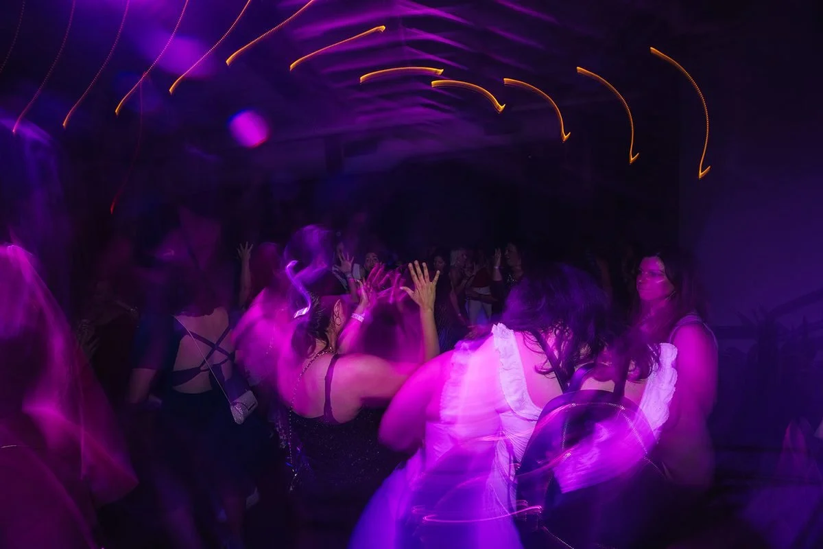 Long-exposure shot of the Dance Her dance floor bathed in purple light with orange light trails on the ceiling and hands raised in the air, Raleigh NC